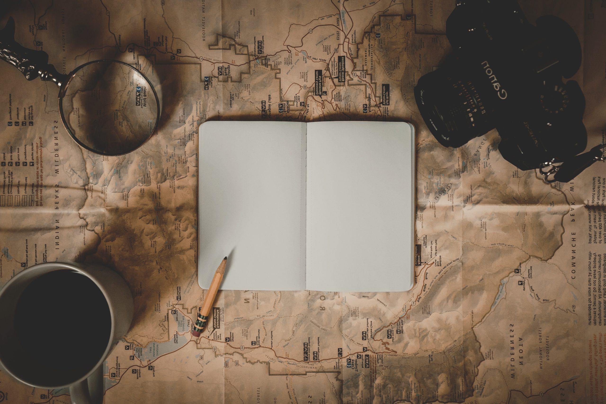 A canon camera, a blank open notebook, a pencil, coffee mug and magnifying glass all sitting on a spread out map