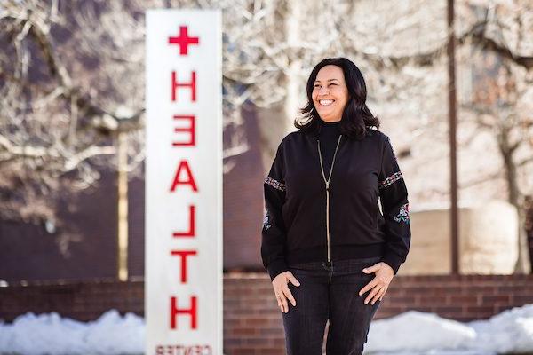 Maria Freyta stands in front of the Health Center at Auraria in a black jacket. The sign behind her reads 'Health'